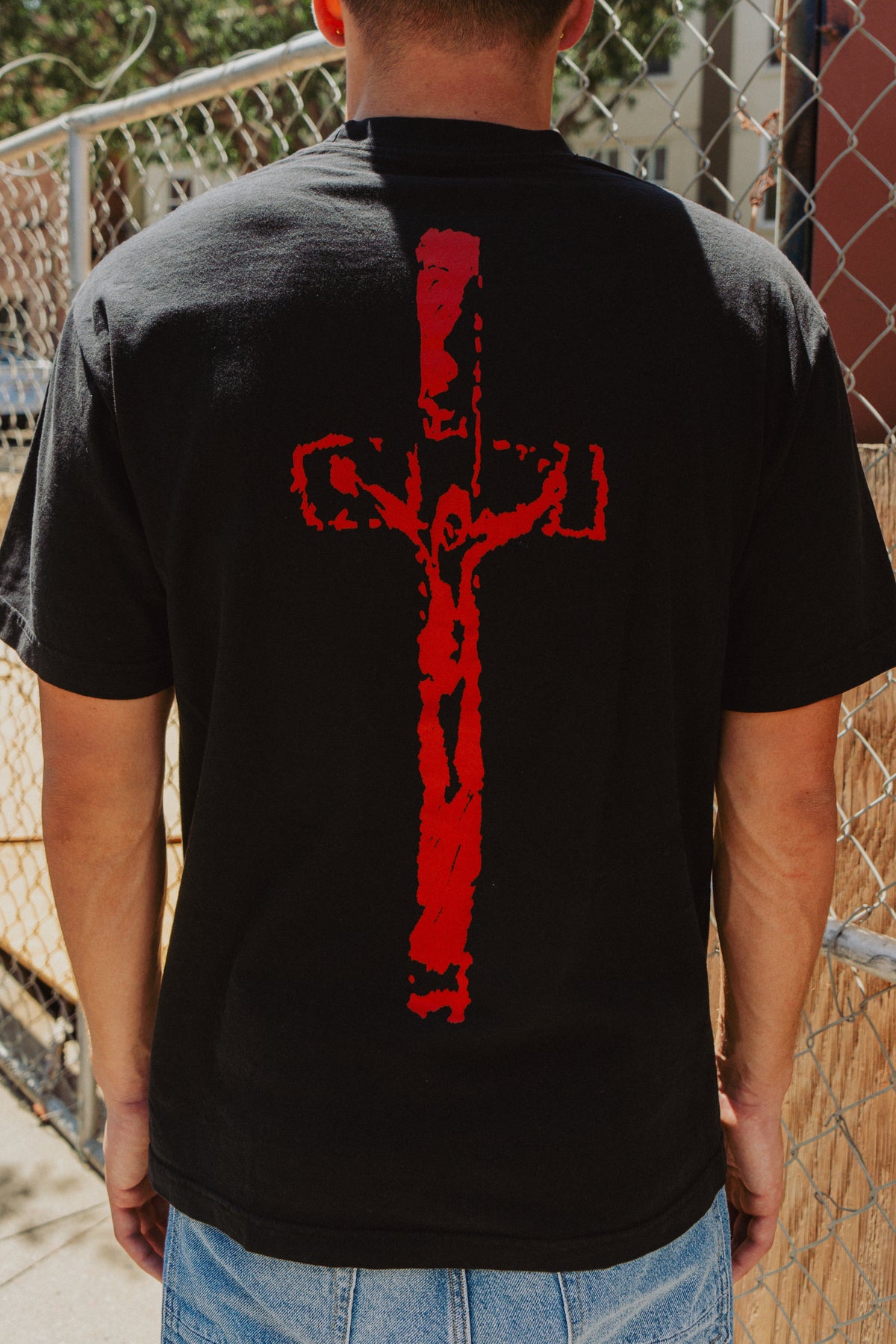 Model wearing black God Is Real tee with red crucifix back graphic by a chain-link fence