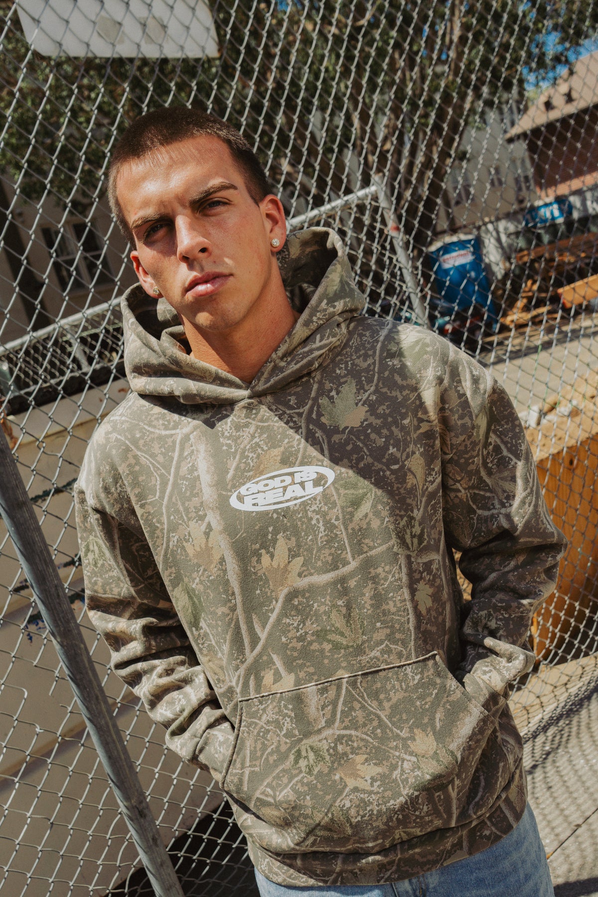 Model wearing camo God Is Real hoodie against a chain-link fence