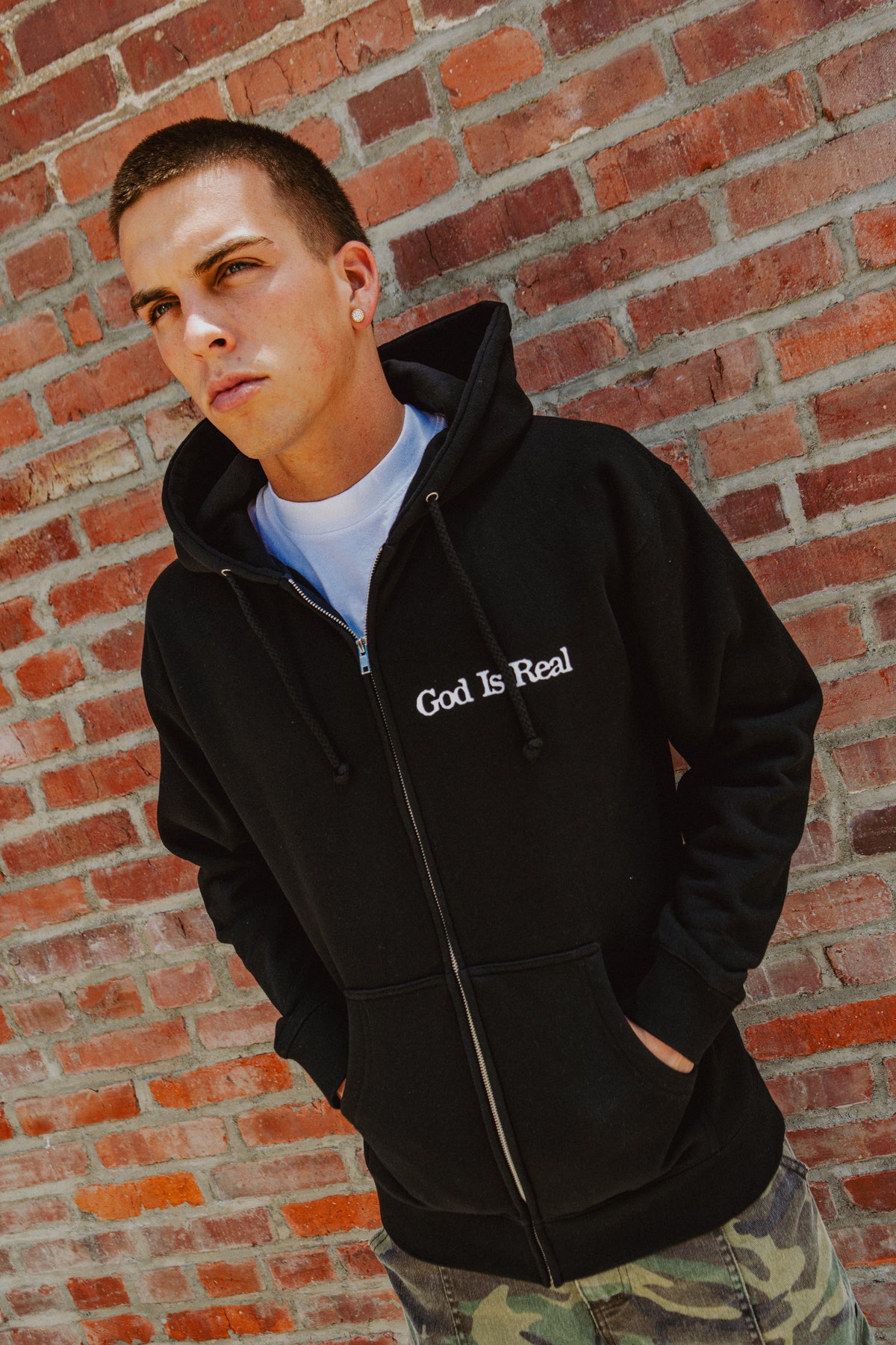 Model wearing black God Is Real Logo Zip Hoodie against a brick wall